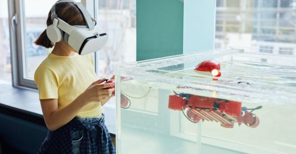 Virtual Reality Developments - Side view of little girl wearing VR goggles and exploring new robot with controllers in light room